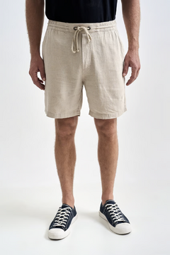 Shorts de linho natural na cor madeira, usados por um homem em pé, com cordão na cintura e tênis escuros.