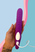 Imagem com mão segurando o Nami Vibrador Strapless com o carregador conectado. Fundo na cor azul com detalhes na cor rosa.