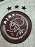 Logotipo do Ajax em uma camisa cinza e bordô, com três estrelas acima do emblema.