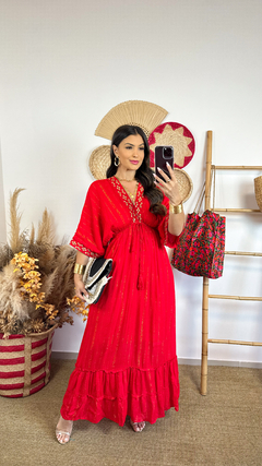 Mulher usando um vestido indiano vermelho com detalhes dourados, segurando uma bolsa preta e posando em frente ao espelho.