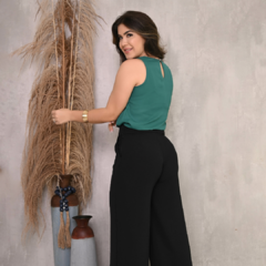 Mulher sorrindo, usando blusa verde e calça preta, posando ao lado de arranjo de plantas secas.