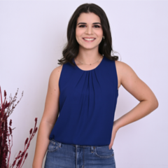 Mulher sorridente usando blusa azul e jeans, posando em um fundo claro com detalhes de plantas.