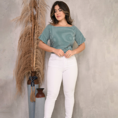 Modelo feminina posando com blusa listrada verde e calça branca, ao fundo decorado com plantas secas e vasos.