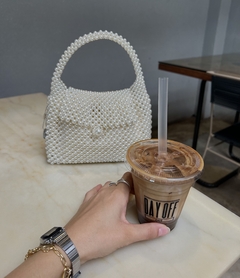 Uma mão segurando um copo de café gelado ao lado de uma bolsa branca decorada com pérolas em uma mesa de mármore.