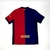 A imagem mostra uma camisa de futebol do FC Barcelona para a temporada 24/25, fabricada pela Nike. A camisa possui listras verticais em azul e vermelho, com o logo da Nike em amarelo no peito direito e o escudo do FC Barcelona no peito esquerdo. O logo do