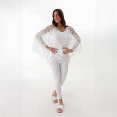 Blusa blanca con detalles bordados, modelo posando de pie con pantalones blancos y sandalias, fondo claro.