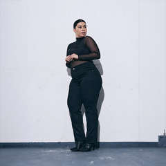 Mujer posando con jeans negros y blusa de manga larga transparente, de pie contra una pared blanca.