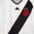 Camisa reserva feminina do Vasco da Gama Kappa 2024-2025. A camisa é branca com a tradicional faixa diagonal preta com contorno vermelho na frente, mangas pretas, gola V com detalhes em preto e branco e textura quadriculada. No lado esquerdo do peito está