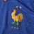 Camisa titular da seleção francesa de futebol 2024-2025. Camisa na cor azul royal com gola V tricolor e o galo, símbolo da seleção, em tamanho maior que o convencional. Na parte interna da gola, a frase "Nos différences nous unissent" (Nossas diferenças n