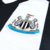 Camisa titular do Newcastle United 2024-25 com listras verticais em preto e branco, gola V preta, detalhes em preto nos ombros e mangas, escudo do NUFC no lado esquerdo do peito, logo da Adidas em preto no lado direito, patrocínio da Sela em branco com bo