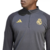 Jaqueta de Treino Real Madrid Adidas UCL 2024 Cinza e Preta. A jaqueta é cinza com detalhes em preto, com o escudo do Real Madrid e o logotipo da Adidas. O modelo está vestindo a jaqueta em um campo de futebol.