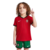 Conjunto Infantil Titular da Seleção Portuguesa Nike 2024-2025. Camisa vermelha com gola preta e verde, calção verde e escudo da FPF. Inspirada na bandeira portuguesa. Vista frontal de um menino vestindo o conjunto completo, em pose de comemoração.