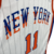 Regata City Edition 2024-25 do New York Knicks. A camisa branca apresenta a icônica marca empilhada "New York" em azul sobre laranja, com detalhes em azul royal nas laterais. O logotipo Nike NYC City Attack aparece no peito.