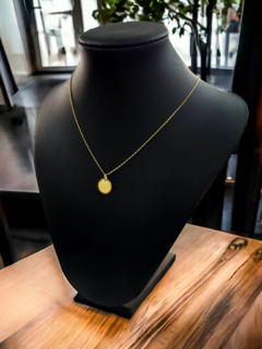 Collar dorado con colgante redondo, exhibido en un maniquí negro sobre una mesa de madera.