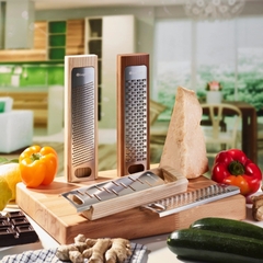 raladores de cozinha, para queijo, chocolate e legumes, feito de madeira e lâmina de aço inox em cima de uma mesa cheia de alimentos