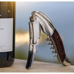 um saca-rolhas de dois estágios de aço inox com acabamento em madeira da marca italiana Legnoart ao lado de uma garrafa de vinho
