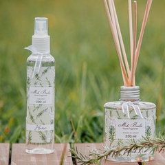 Frasco transparente de spray para ambientes de aroma Alecrim, da marca Mels Brushes, produto ladeado de frasco de difusor para ambientes, ao fundo gramado verde.