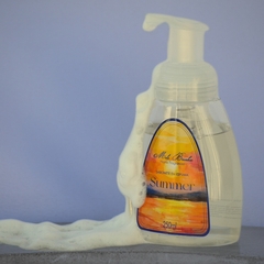 Frasco transparente de sabonete líquido em espuma aroma Autunn, da marca Mels Brushes, imagem com espuma saindo do frasco ambientada com fundo azul.