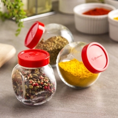 três potes de vidro com tampa vermelha com temperos e condimentos variados dentro em cima de uma mesa
