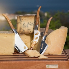 um kit de 3 facas de queijo, contendo um modelo para queijo duro, um modelo para queijo parmesão e um modelo para queijo mole, com lâmina de aço inox e cabo de madeira da marca Legnoart, sobre uma mesa com pedaços de queijo