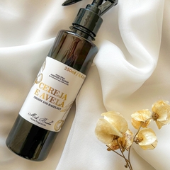Home Spray e Tecidos com frasco âmbar e rótulo branco e dourado, aroma cereja e avelã, da marca Mels Brushes, sobre fundo com tecido perolado e flores amarelas.