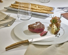 Um prato branco com um pedaço de carne ao lado de uma faca de aço japones com cabo de madeira da marca Legnoart