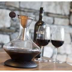 um decanter de vidro, em cima de uma base de madeira, com um funil de aço inox e duas taças de vinho tinto sobre uma mesa