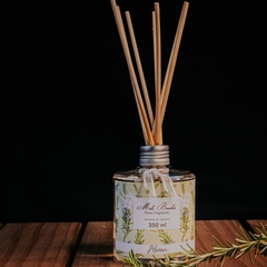 Frasco transparente de difusor para ambientes de aroma Alecrim com varetas, da marca Mels Brushes, imagem com fundo preto e produto sobre mesa de madeira.