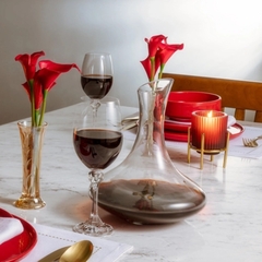 Uma mesa branca com um vaso cheio de flores vermelhas, com um decanter para vinhos de vidro e duas taças cheia de vinho