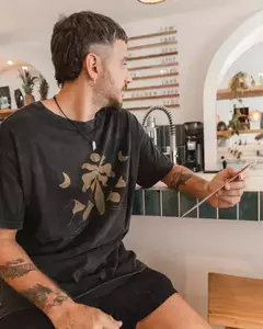 Hombre con remerón negro de la colección 2023, sentado en un café, sosteniendo un menú y con decoración de plantas en el fondo.