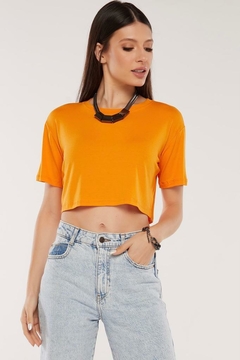 Modelo morena usando um cropped liso feminino laranja em viscolycra atacado, cabeça levemente virada para a direita, olhando para baixo com a mão direita no bolso, sobre um fundo branco. G&B Bros