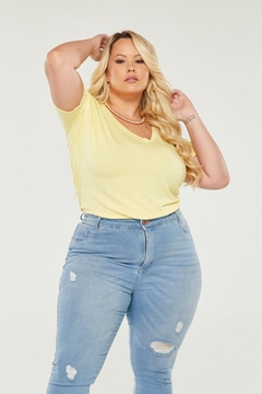 Modelo plus size loira vestindo uma camiseta amarela de gola v e viscolycra, olhando para a câmera com os braços erguidos na altura da cabeça, sobre um fundo branco.