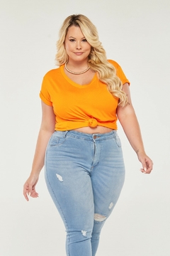 Modelo plus size loira vestindo uma camiseta laranja de gola v e viscolycra com nó, olhando para a câmera com um leve sorriso, sobre um fundo branco.