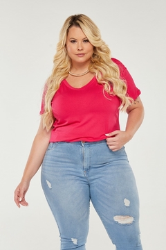 Modelo plus size loira vestindo uma camiseta pink de gola v e viscolycra, olhando para a câmera, segurando a barra da camiseta com a mão esquerda, sobre um fundo branco.