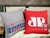 Duas almofadas promocionais repousam sobre uma cama. A almofada à esquerda é cinza e apresenta o logo e o skyline em azul e vermelho da Educador AM 1060 de Piracicaba. A almofada à direita é vermelha e exibe o logo em branco da JP FM Piracicaba-103.1.