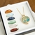 Imagem colorida mostra corrente veneziana, pingente relicário árvore da vida dourado e 7 pedras naturais. O colar e as pedras estão posicionados dentro de uma caixa pequena de jóias. 