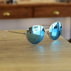 anteojos de sol de metal de forma redonda color plata satinada con lentes polarizados espejados color celeste modelo los pioneros marcca nomade con fondo de madera