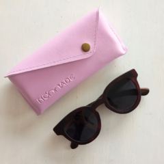 estuche para anteojos de cuero sintetico color lila con cierre de boton marca Nomade al lado de anteojos de sol de forma redondeada fondo blanco