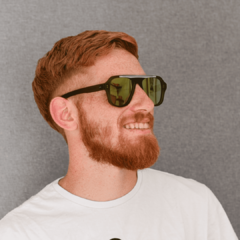 hombre joven de piel blanca, pelo y barba de color rojizo sonriendo con anteojos de sol de acetato color verde con lentes espejados color bronce modelo Patagonia PA marca Nomade con fondo gris