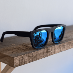 anteojos de sol de acetato color negro mate con lentes espejados polarizados color azul modelo Tulum PA marca Nómade sobre fondo de madera y blanco