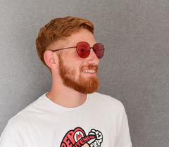 hombre joven de piel blanca y pelo rojo con anteojos de sol de metal estilo redondo color cobre con lentes color rosa modelo Los Pioneros marca Nomade con fondo gris