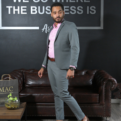 Hombre posando en un traje gris con camisa rosa, frente a un fondo oscuro y un sofá de cuero marrón.