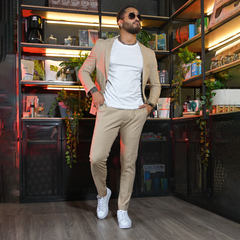 Hombre elegante vestido con un traje beige y camiseta blanca, posando en un ambiente moderno con estanterías y plantas.